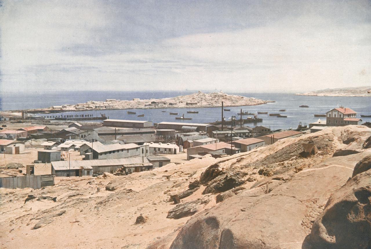 Historische Aufnahme von Lüderitz mit Gebäuden in Küstennähe und der langgestreckten Haifischinsel im Hintergrund. Historische Aufnahme von Lüderitz mit Gebäuden in Küstennähe und der langgestreckten Haifischinsel im Hintergrund.