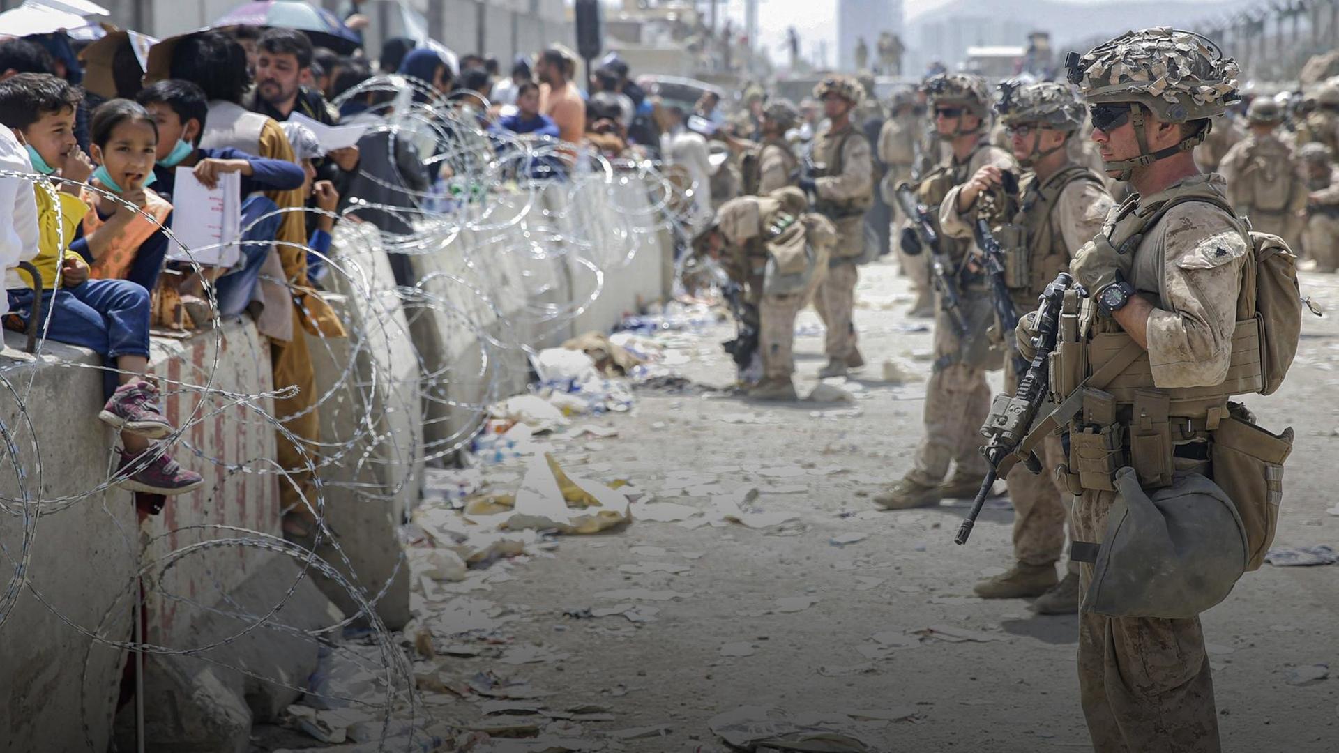 Bewaffnete Soldaten sichern den Flughafen Kabul in Afghanistan 2021, vor den Mauern stehen zahlreiche Menschen und Kinder hinter Stacheldraht. Bewaffnete Soldaten sichern den Flughafen Kabul in Afghanistan 2021, vor den Mauern stehen zahlreiche Menschen und Kinder hinter Stacheldraht.