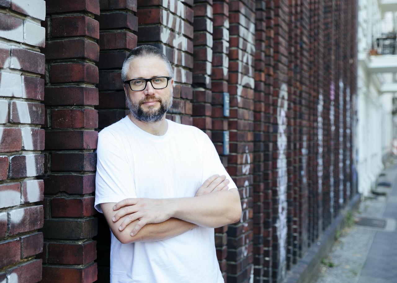 Der Journalist und Buchautor Daniel Schulz lehnt im weißen Shirt an einer Mauer. Er trägt Brille und Bart.
