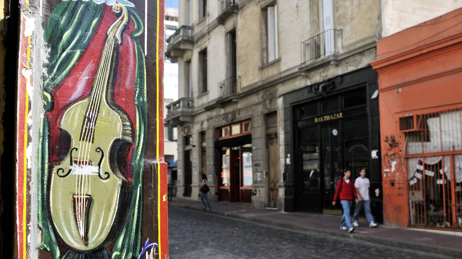 Zwei Menschen gehen im historischer Stadtteil San Telmo in Buenos Aires an einer Hauswand vorbei, die mit einer Geige bemalt ist und dabei von einem grünen Vorhang umgeben ist. Zwei Menschen gehen im historischer Stadtteil San Telmo in Buenos Aires an einer Hauswand vorbei, die mit einer Geige bemalt ist und dabei von einem grünen Vorhang umgeben ist.