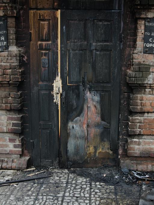 Eine verbrannte Haustür in einem kaputten Haus. Davor steht ein Stuhl.
