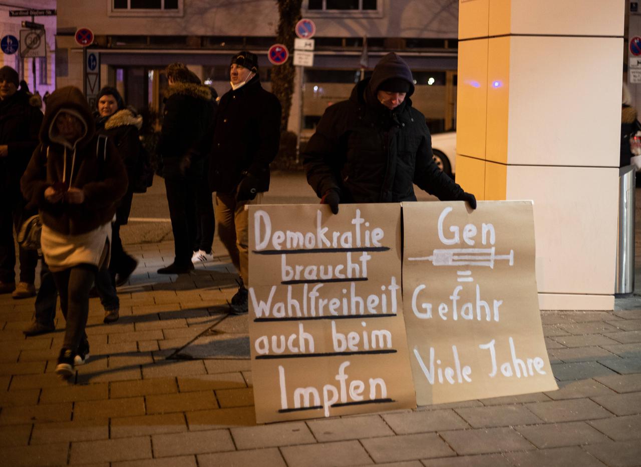 Auf einem Schild steht: "Demokratie braucht Wahlfreiheit auch beim Impfen", auf einem zweiten "Gen = Gefahr Viele Jahre". Am 5. Januar 2022 riefen verschiedene Querdenken-Gruppen zu illegalen Versammlungen, getarnt als Spaziergänge, in München auf. 