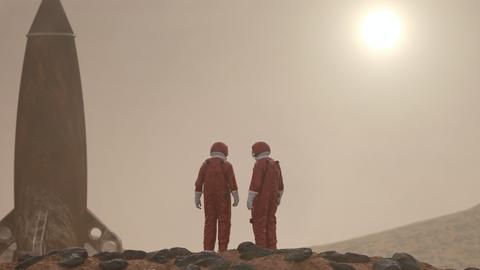 Zwei Personen in roten Ganzkörperanzügen auf rotem, steinigen Untergrund. Neben ihnen eine Rakete, aufrecht stehend. Am Horizont die Sonne.