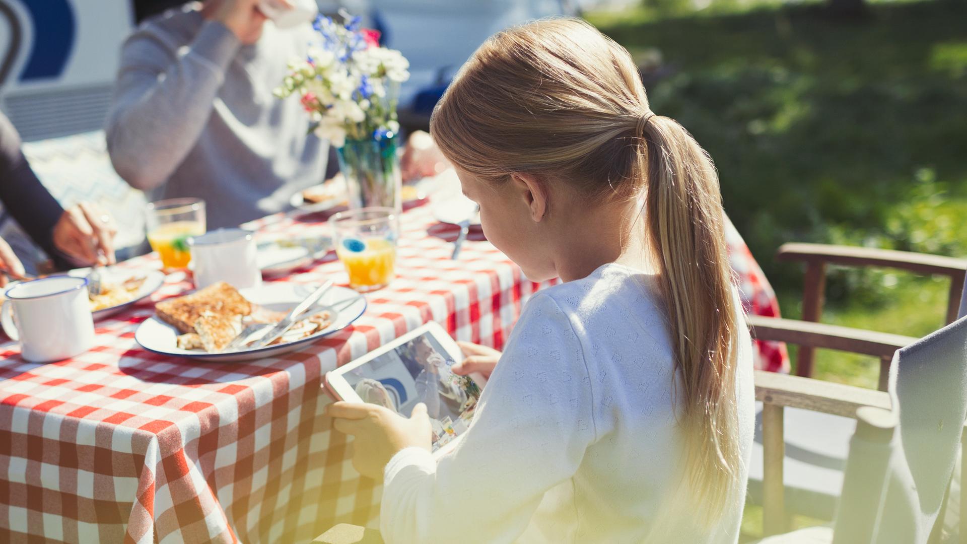 Ein Mädchen schaut auf das Display eines Tablets, während sie draußen, mit Ihrer Familie beim Camping frühstückt.