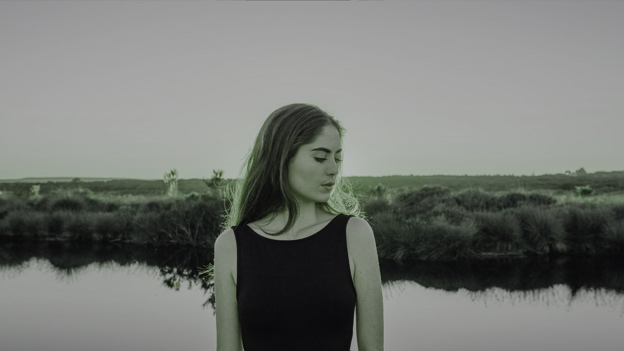 Eine junge Frau, mit dem Kopf zur Seite gesenkt, steht vor einem See. Im Hintergrund ist eine Wiesenlandschaft.