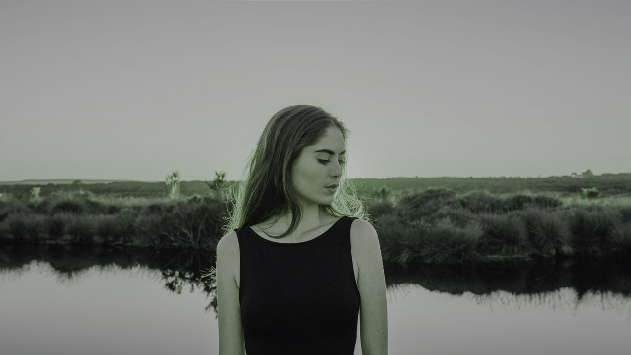 Eine junge Frau, mit dem Kopf zur Seite gesenkt, steht vor einem See. Im Hintergrund ist eine Wiesenlandschaft. Eine junge Frau, mit dem Kopf zur Seite gesenkt, steht vor einem See. Im Hintergrund ist eine Wiesenlandschaft.