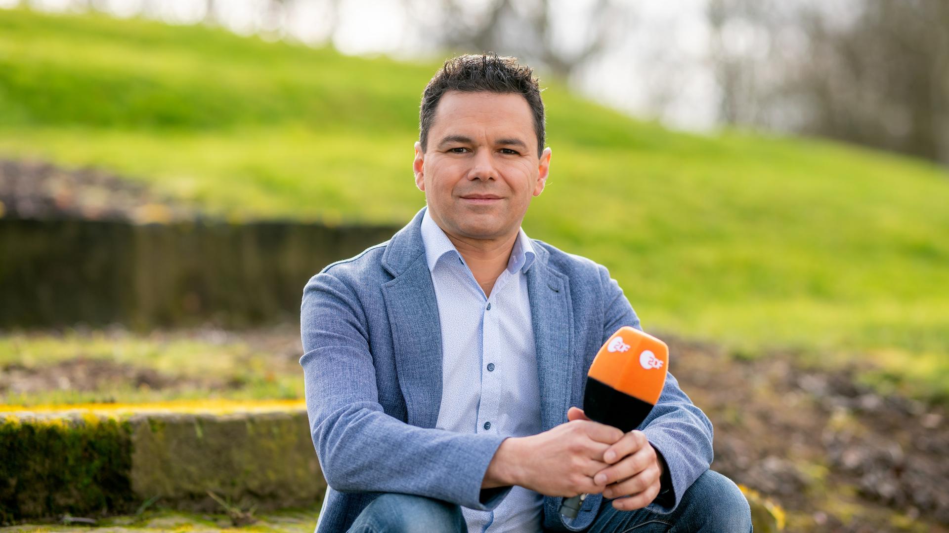 Ein Mann mit braunem Haar und blauem Anzug hält ein ZDF-Mikro in der Hand und blickt in die Kamera. Ein Mann mit braunem Haar und blauem Anzug hält ein ZDF-Mikro in der Hand und blickt in die Kamera.
