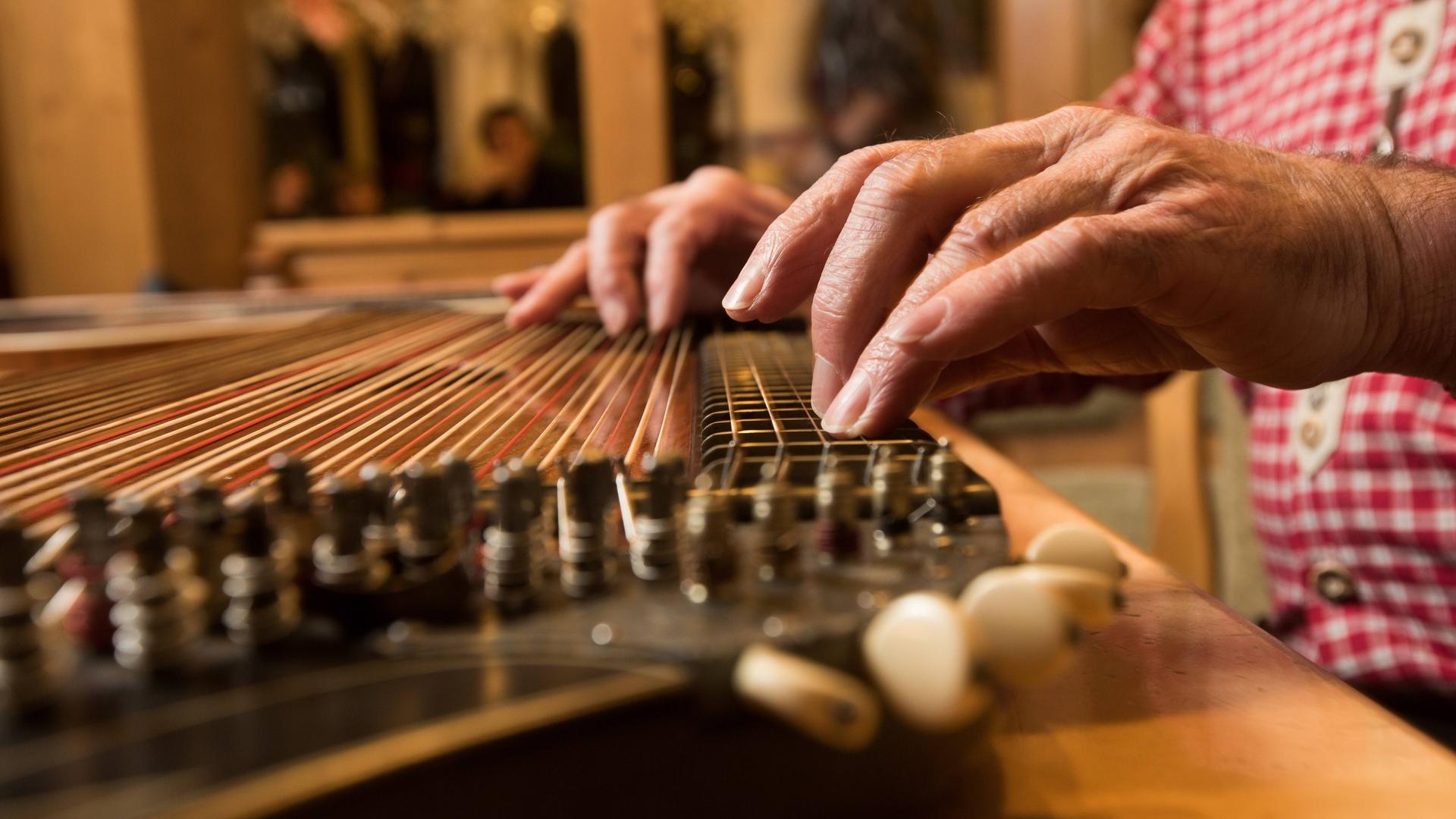 Auf den Saiten einer Zither spielen zwei ältere Hände, die zu einem Mann gehören, der in Tracht spielt. Auf den Saiten einer Zither spielen zwei ältere Hände, die zu einem Mann gehören, der in Tracht spielt.