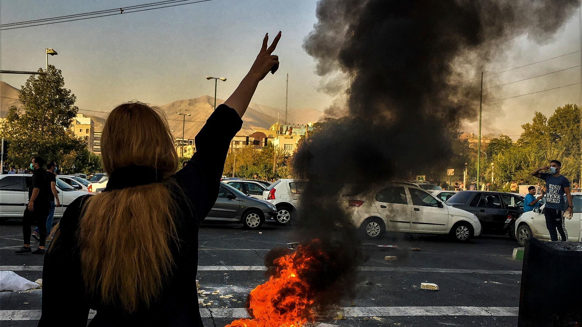 Eine Frau steht während einer Demonstration vor einem brennenden Auto-Reifen. Sie zeigt das Sieges-Zeichen.