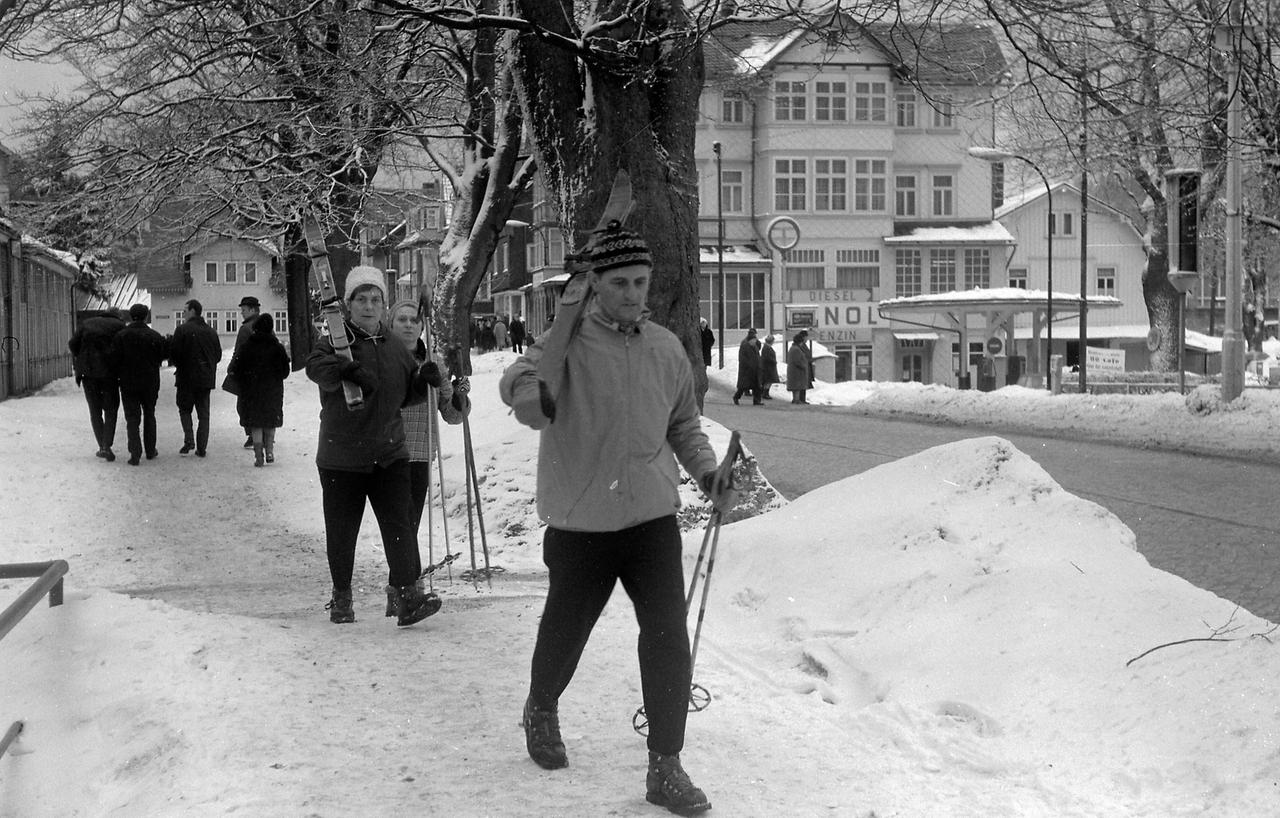 Oberhof Winter 1970 Historisches Bild und Foto aus Oberhof im Thüringer Wald. Zu sehen Skiläufer, Wintersportler die eine verschneite Straße entlang gehen. Schnee türmt sich am Straßenrand. Im Hintergrund Tankstelle Minol. Historical Image and Photography from snowy Oberhof Thuringia 