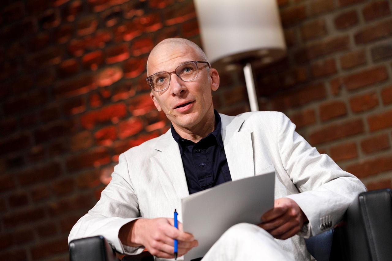 Armen Avanessian in einem weissen Anzug, sitzt in einem Ledersessel und spricht auf der phil.COLOGNE. Armen Avanessian in einem weissen Anzug, sitzt in einem Ledersessel und spricht auf der phil.COLOGNE.