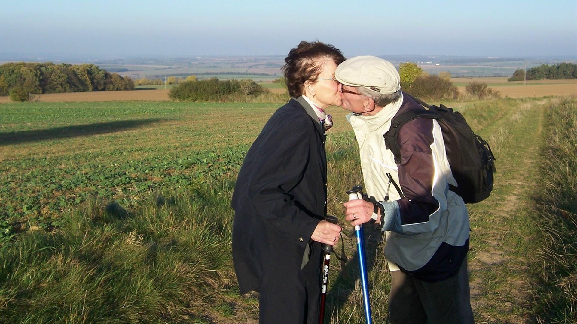 Ein älteres Paar steht mit Nordic-Walking-Stöcken küssend auf einem Feld. Ein älteres Paar steht mit Nordic-Walking-Stöcken küssend auf einem Feld.