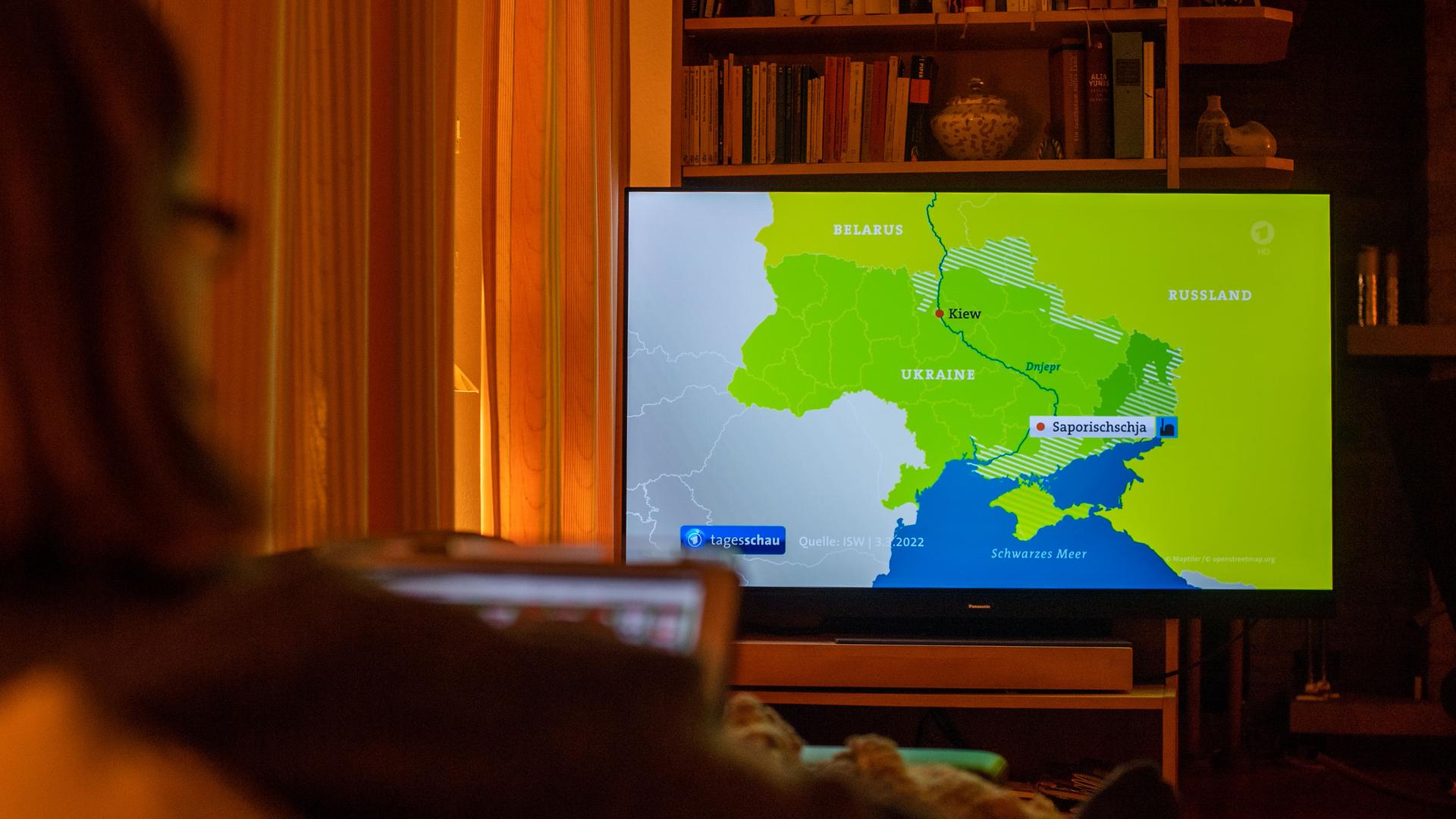 Eine Frau sitzt auf dem Sofa und schaut im Fernsehen die Tagesschau zum Ukraine-Konflikt. Eine Frau sitzt auf dem Sofa und schaut im Fernsehen die Tagesschau zum Ukraine-Konflikt.