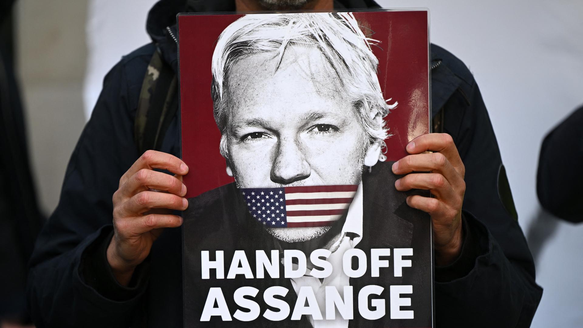 Ein Aktivist hält ein Plakat von WikiLeaks Gründer Julian Assange, der den Mund mit einer amerikanischen Flagge zugeklebt hat. Der Text darauf lautet: "Hands off Assange. Don't shoot the Messenger", London, April 2022.