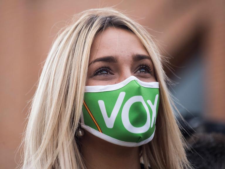 Eine junge blonde Frau trägt eine grüne Maske, auf der mit weißen Buchstaben Vox steht.