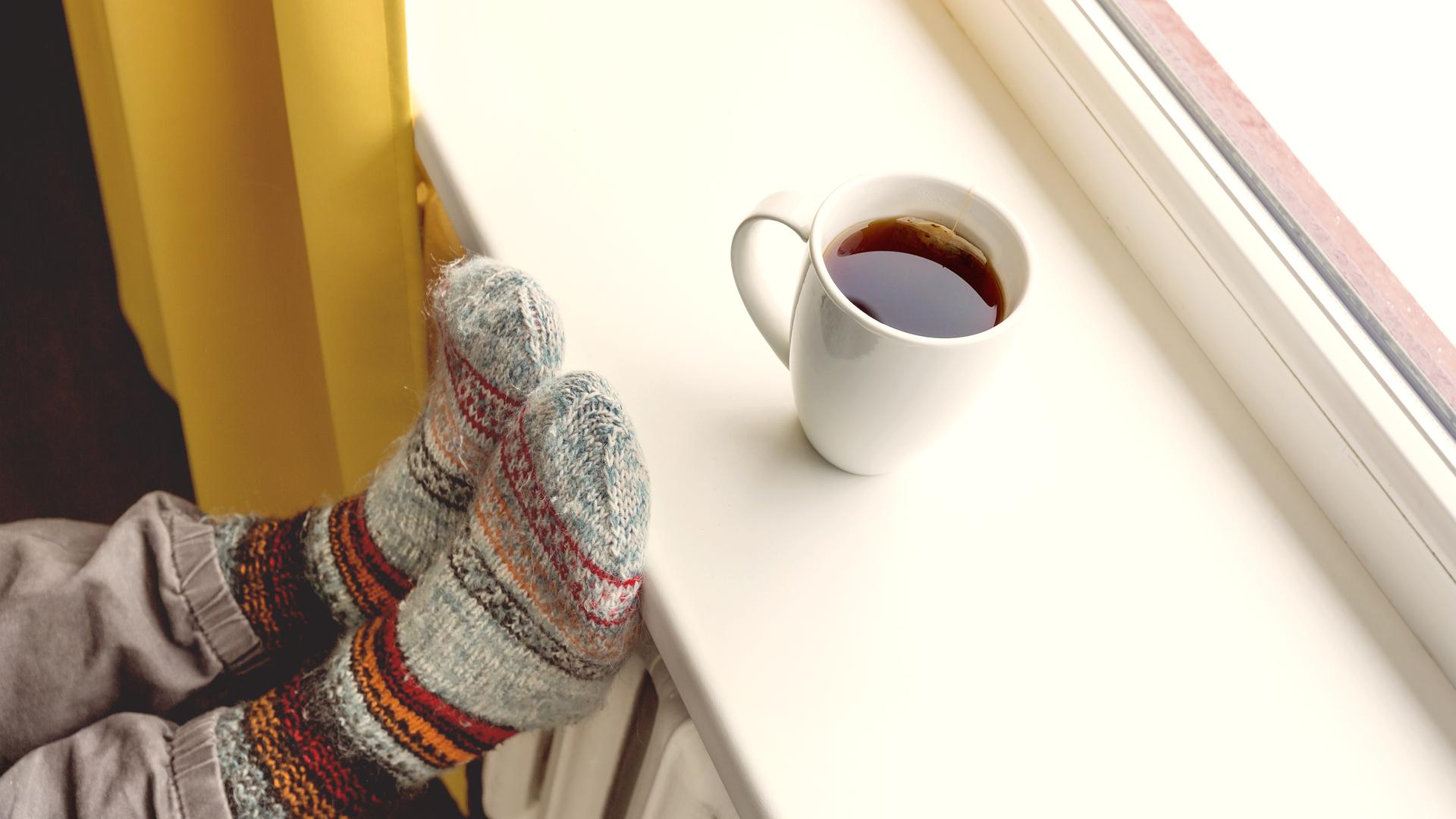 Eine Tasse mit heissem Tee steht auf einer Fensterbank, Füße in Wollsocken liegen auf der Heizung. Eine Tasse mit heissem Tee steht auf einer Fensterbank, Füße in Wollsocken liegen auf der Heizung.