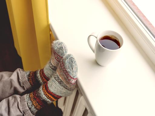 Eine Tasse mit heissem Tee steht auf einer Fensterbank, Füße in Wollsocken liegen auf der Heizung.