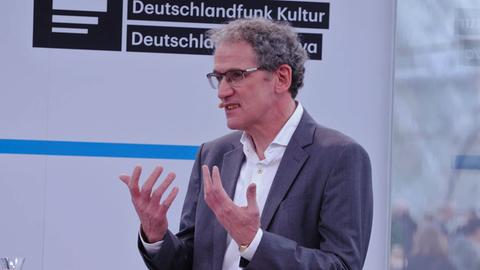 Dirk Oschmann in weissem Hemd und grauer Jacke spricht vor einer weissen Wand mit Deutschlandradio Logos im Hintergrund.