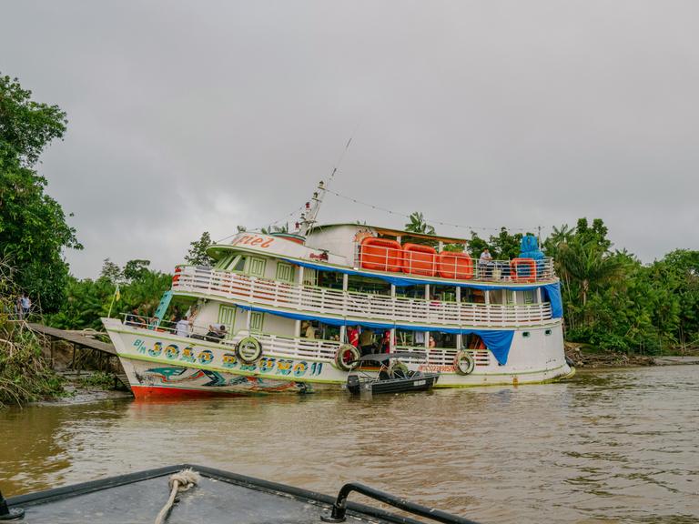 Ein zweitstöckiges Schiff liegt am Ufer eines großen tropischen Flusses, über einen Steg gehen Menschen an Bord