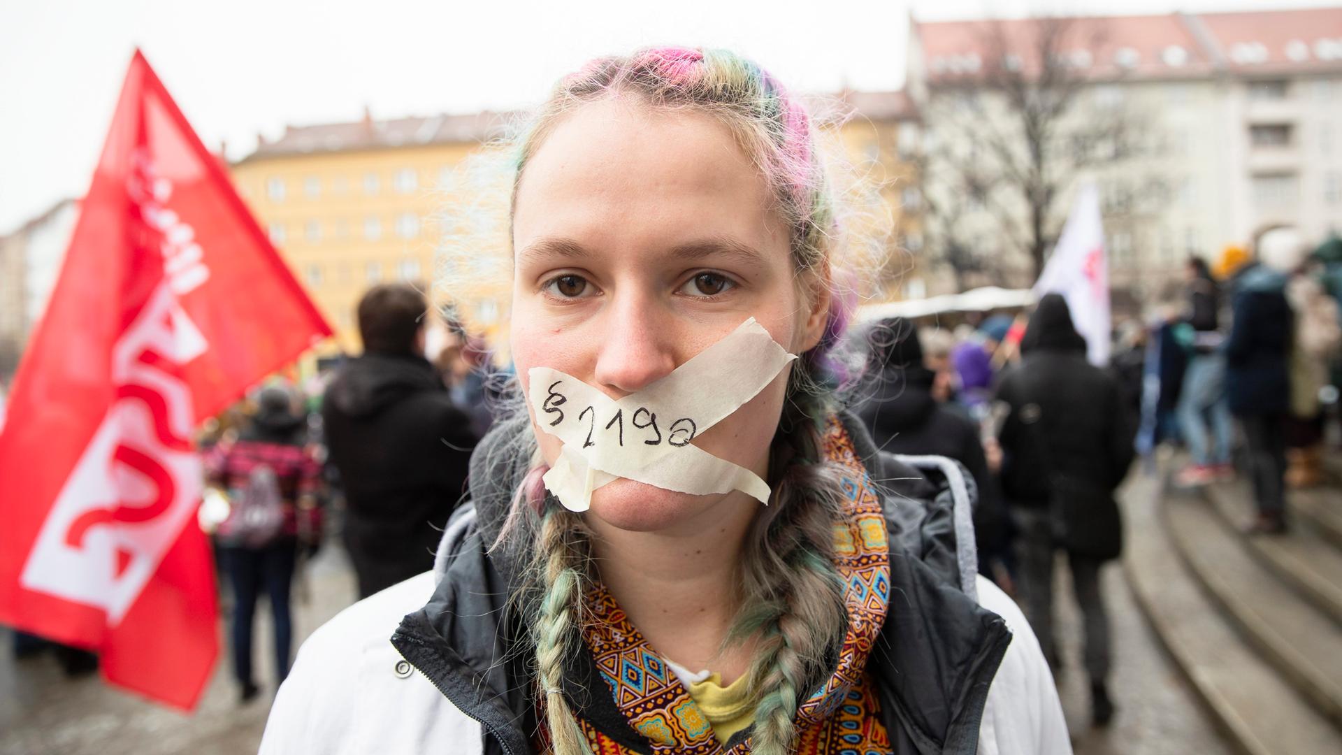 Eine junge Frau bei einer Demo. Sie hat mit Klebeband ein Kreuz über den Mund geklebt, das es ihr unmöglich macht zu sprechen. Auf dem Klebeband steht "Paragraf 219a" Eine junge Frau bei einer Demo. Sie hat mit Klebeband ein Kreuz über den Mund geklebt, das es ihr unmöglich macht zu sprechen. Auf dem Klebeband steht "Paragraf 219a"