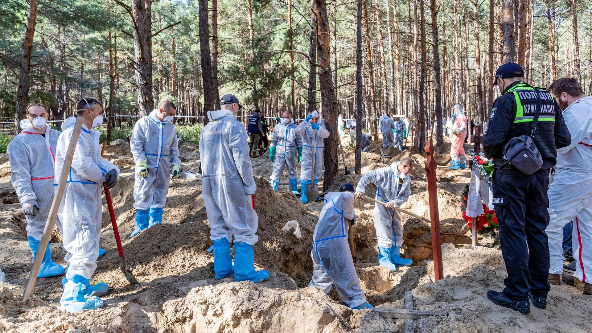 Zahlreiche Forensiker stehen in weißen Schutzanzügen, mit Mund-Nasen-Schutz und Schaufeln in einem Waldstück. Der sandige Boden ist an mehreren Stellen ausgehoben. Zahlreiche Forensiker stehen in weißen Schutzanzügen, mit Mund-Nasen-Schutz und Schaufeln in einem Waldstück. Der sandige Boden ist an mehreren Stellen ausgehoben.