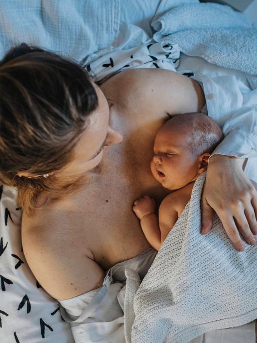 Vogelperspektive auf eine blonde Frau, die mit einem Neugeborenen im Arm im Bett liegt.
