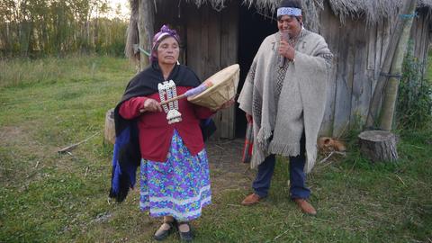 Eine Frau in traditioneller Kleidung und großem Silberschmuck am Hals spielt eine Trommel, während ein mann, ebenfalls mit traditionellem Umhang, ein Blasinstrument spielt.