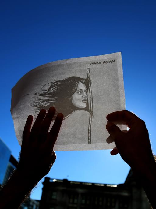 Zeichnung von Mahsa Amini, deren Haare eine Flagge bilden, auf einem Plakat, das während eines Protests hochgehalten wird.