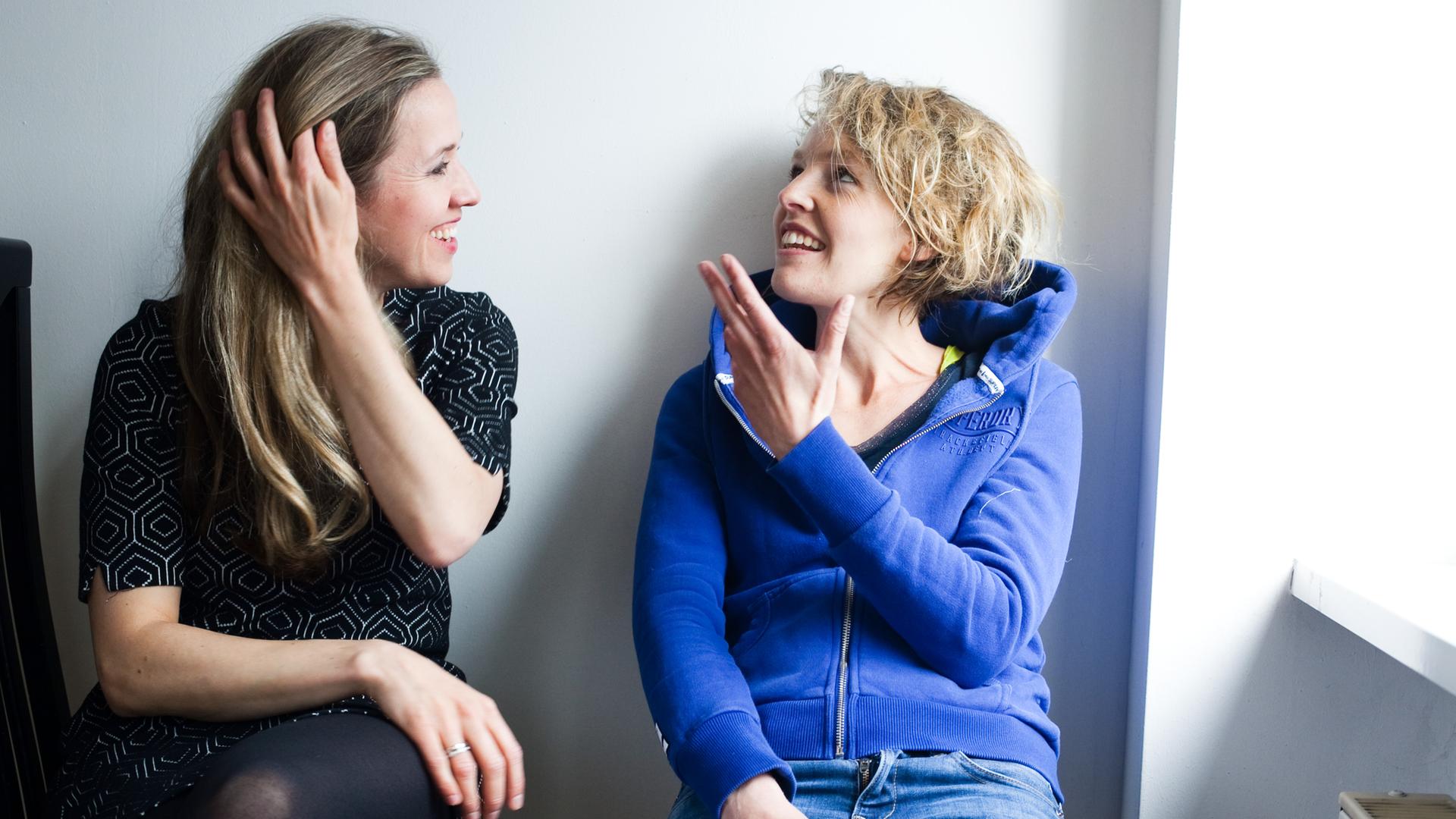 Zwei junge Frauen sitzen vor einer Wand und unterhalten sich. Die Frau links im Bild, Annika Sophie Mendrala, hört lächelnd zu. Verena Usemann, die Frau rechts, gestikuliert beim Sprechen und schaut leicht nach oben. Zwei junge Frauen sitzen vor einer Wand und unterhalten sich. Die Frau links im Bild, Annika Sophie Mendrala, hört lächelnd zu. Verena Usemann, die Frau rechts, gestikuliert beim Sprechen und schaut leicht nach oben.