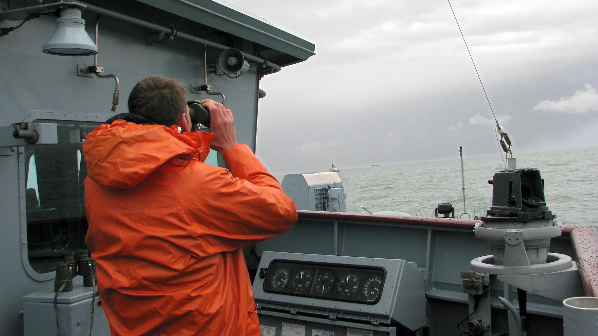 Ein Marinesoldat in Regenjacke steht als Ausguck auf einem Schiff und betrachtet das Meer durch ein Fernglas.