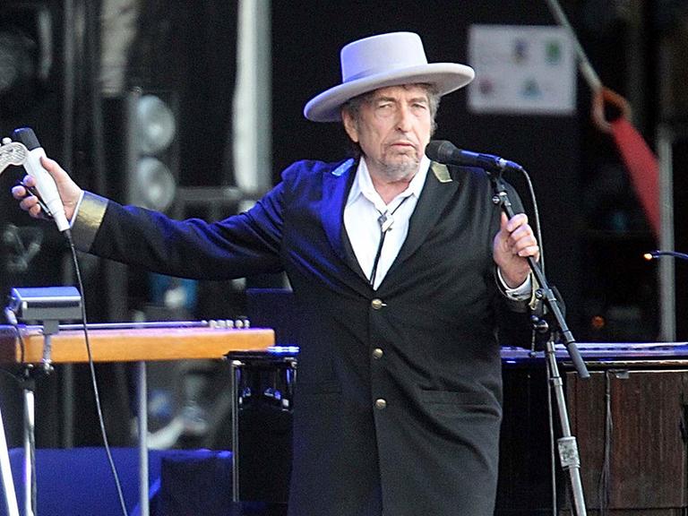 Ein Mann steht in Anzug und Hut auf der Bühne und hält ein Mikrofon in jeder Hand.