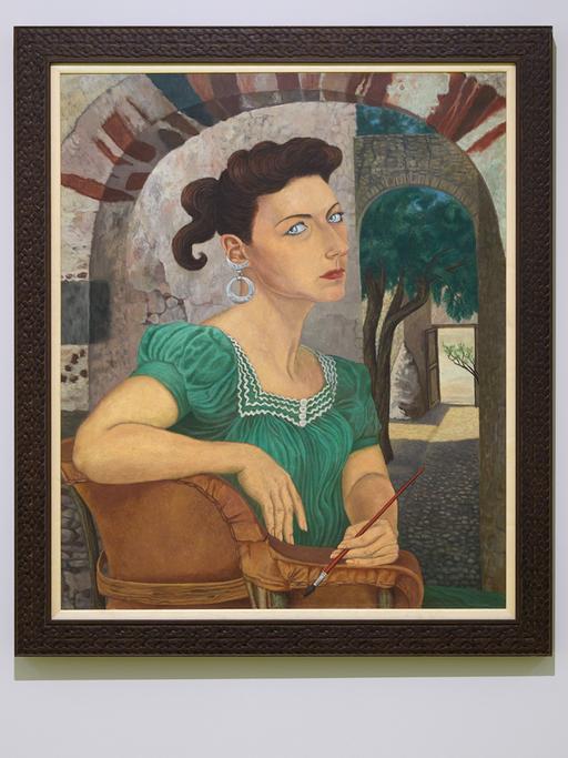 Das Selbstporträt der Malerin Olga Costa zeigt eine dunkelhaarige Frau Mitte 30 im grünen Sommerkleid sitzend unter freiem Himmel in einem Hof. Sie hält einen kleinen Malpinsel in der linken Hand und fixiert den Betrachter mit ernstem Blick.
