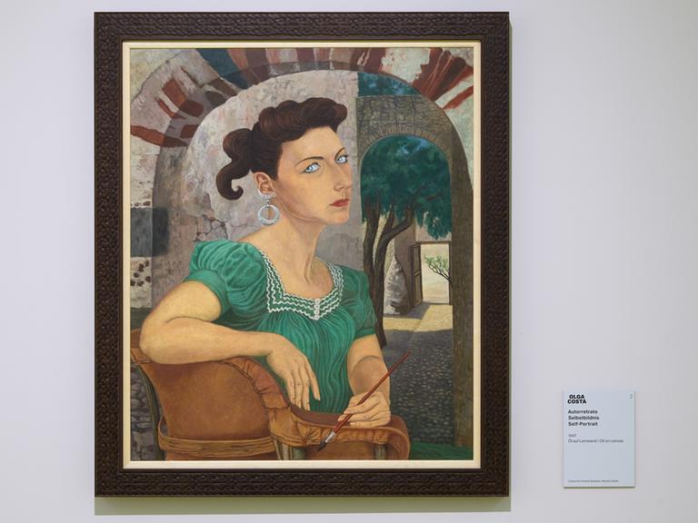 Das Selbstporträt der Malerin Olga Costa zeigt eine dunkelhaarige Frau Mitte 30 im grünen Sommerkleid sitzend unter freiem Himmel in einem Hof. Sie hält einen kleinen Malpinsel in der linken Hand und fixiert den Betrachter mit ernstem Blick.