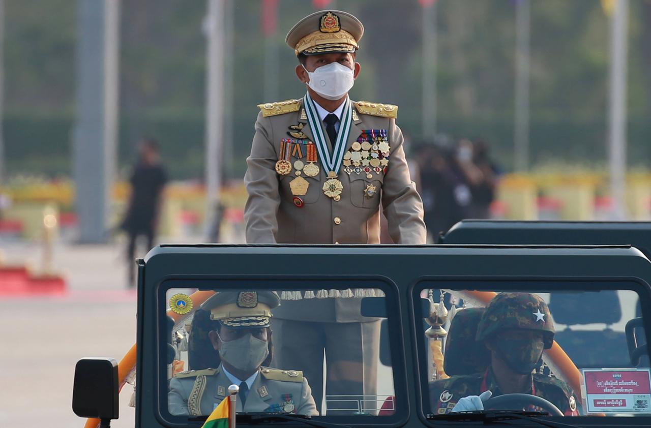 Ein mit vielen goldenen Orden behängter Militär in Uniform und mit Mundschutz steht hinten auf einem fahrenden Jeep.