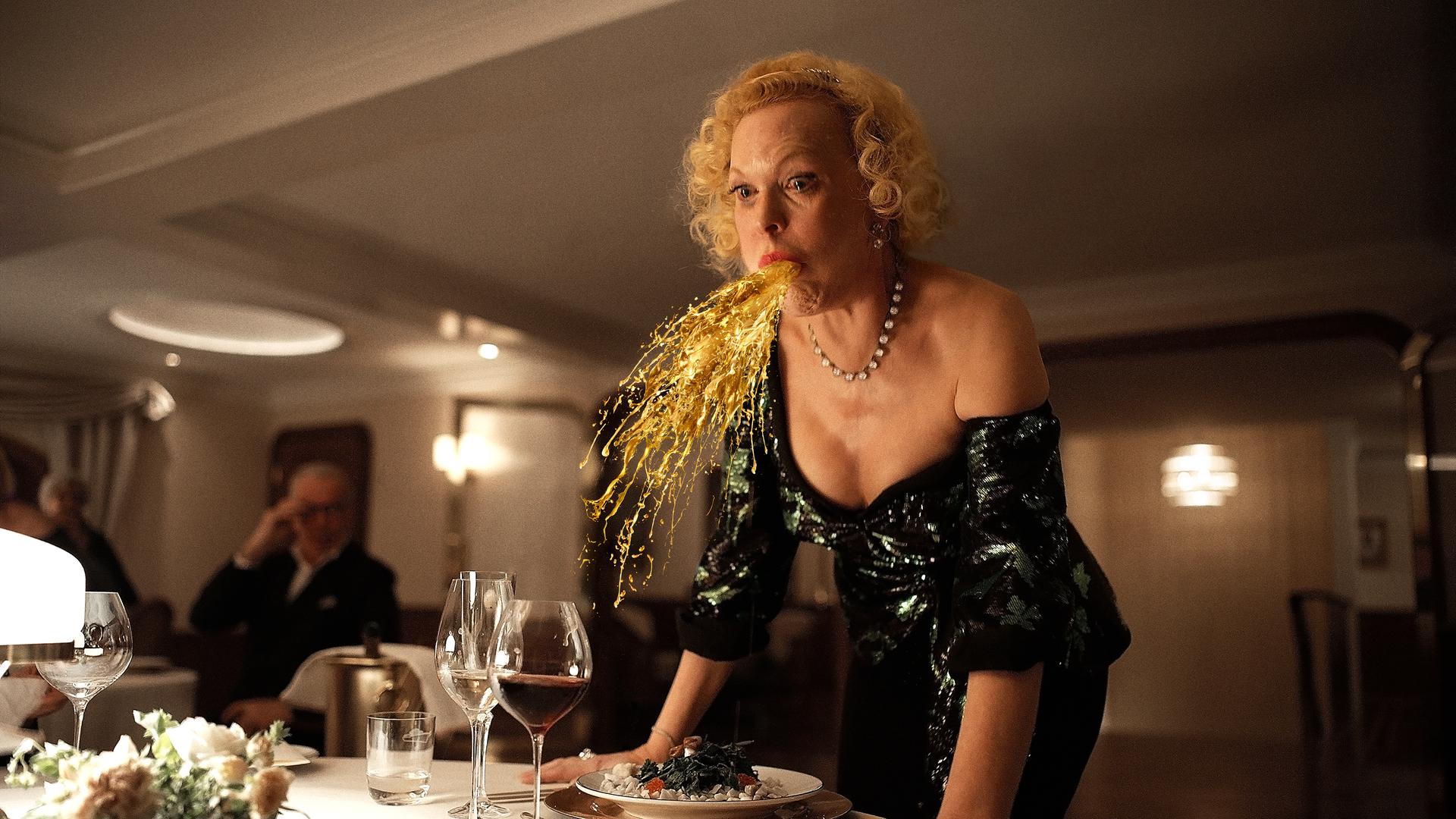 Der Film "Triangle of Sadness", bei den Oscars 2023 nominiert: Eine Frau speiht eine Art Goldregen an einem festlichen gedeckten Tisch. Der Film "Triangle of Sadness", bei den Oscars 2023 nominiert: Eine Frau speiht eine Art Goldregen an einem festlichen gedeckten Tisch.