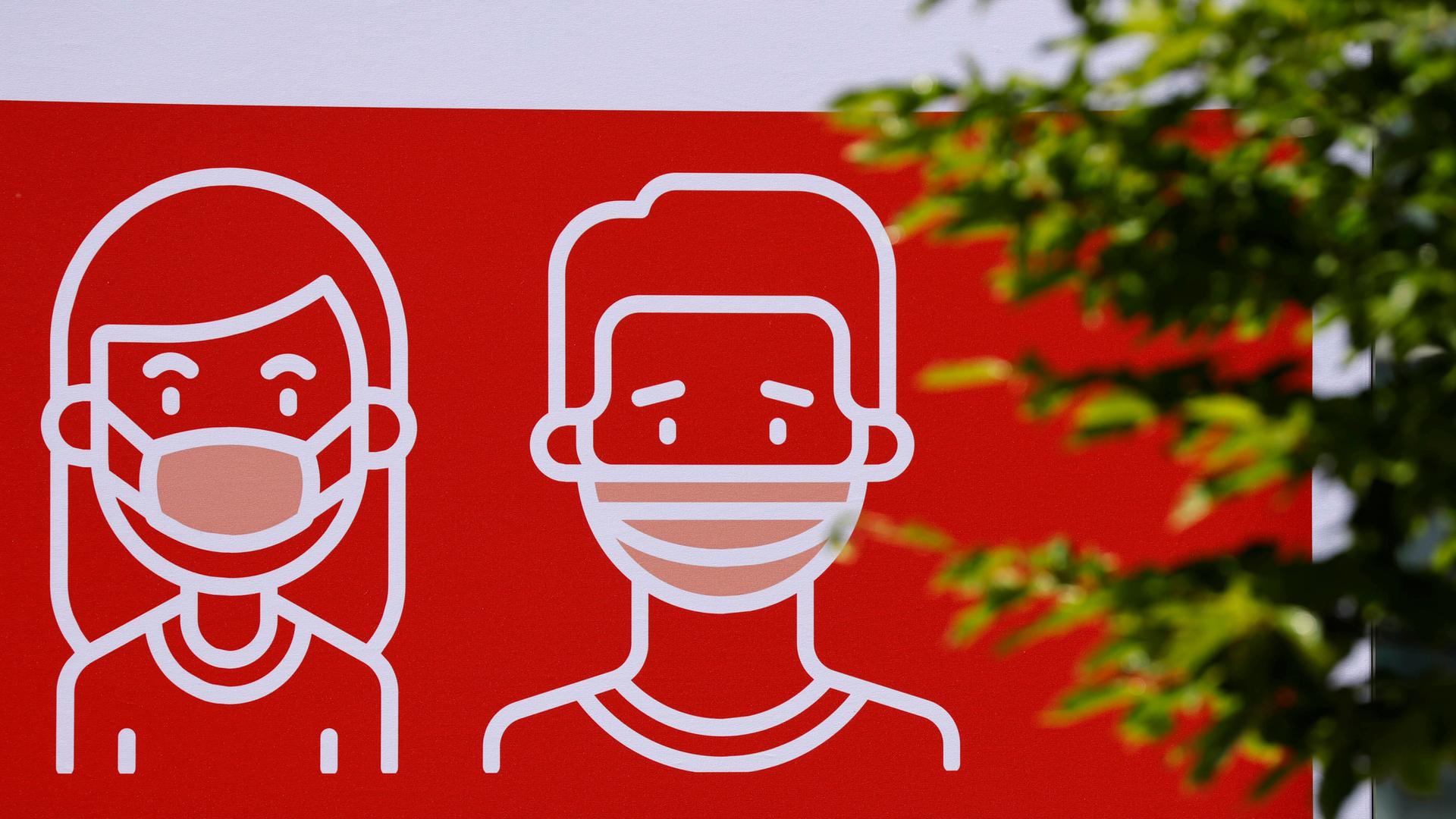 Auf einem roten Schild sind zwei Menschen gezeichnet. Sie tragen eine Maske. 