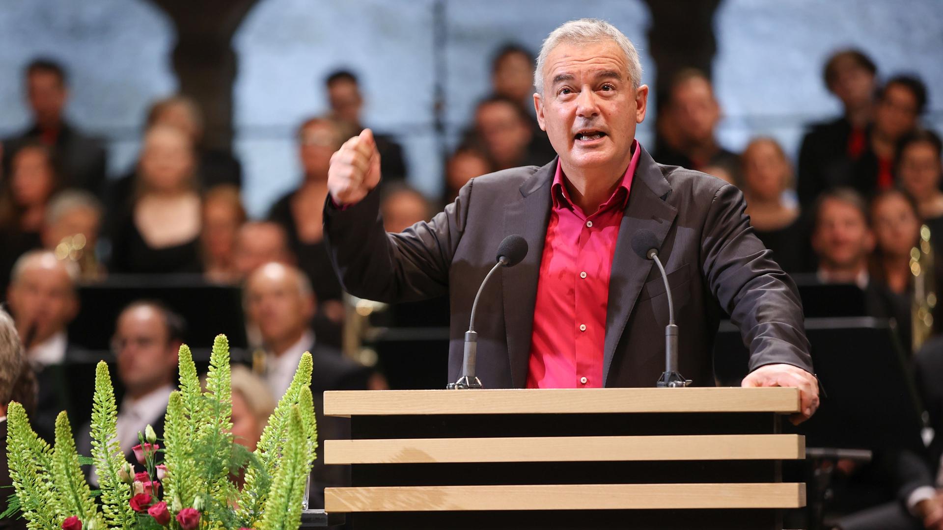 Der Schriftsteller Ilija Trojanow, am Rednerpult in schwarzem Sakko und rotem Hemd, hat die rechte Hand erhoben, um einer Aussage Nachdruck zu verleihen. Der Schriftsteller Ilija Trojanow, am Rednerpult in schwarzem Sakko und rotem Hemd, hat die rechte Hand erhoben, um einer Aussage Nachdruck zu verleihen.