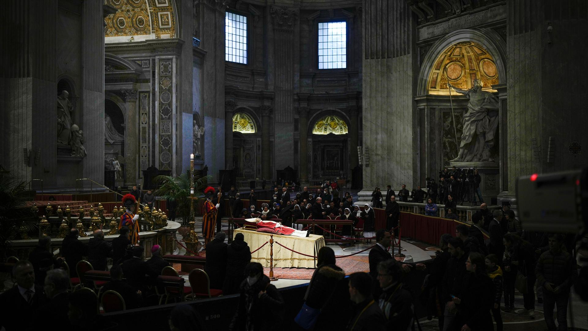 Der emeritierte Papst liegt aufgebahrt im Petersdom. Um ihn versammelt stehen und knien betende Menschen. Der emeritierte Papst liegt aufgebahrt im Petersdom. Um ihn versammelt stehen und knien betende Menschen.