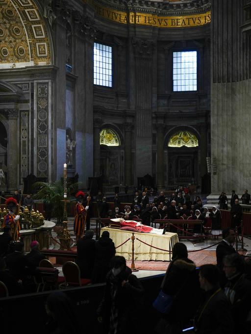 Der emeritierte Papst liegt aufgebahrt im Petersdom. Um ihn versammelt stehen und knien betende Menschen.