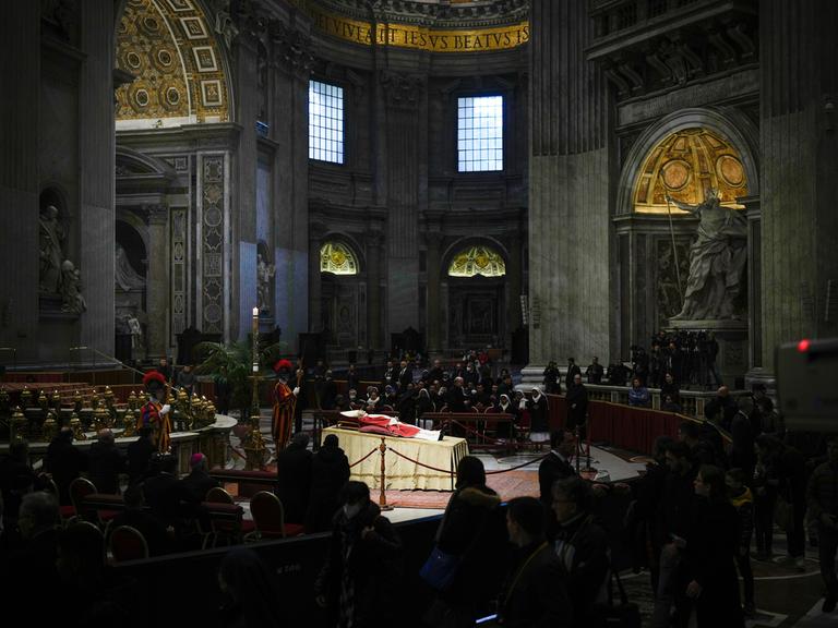 Der emeritierte Papst liegt aufgebahrt im Petersdom. Um ihn versammelt stehen und knien betende Menschen.