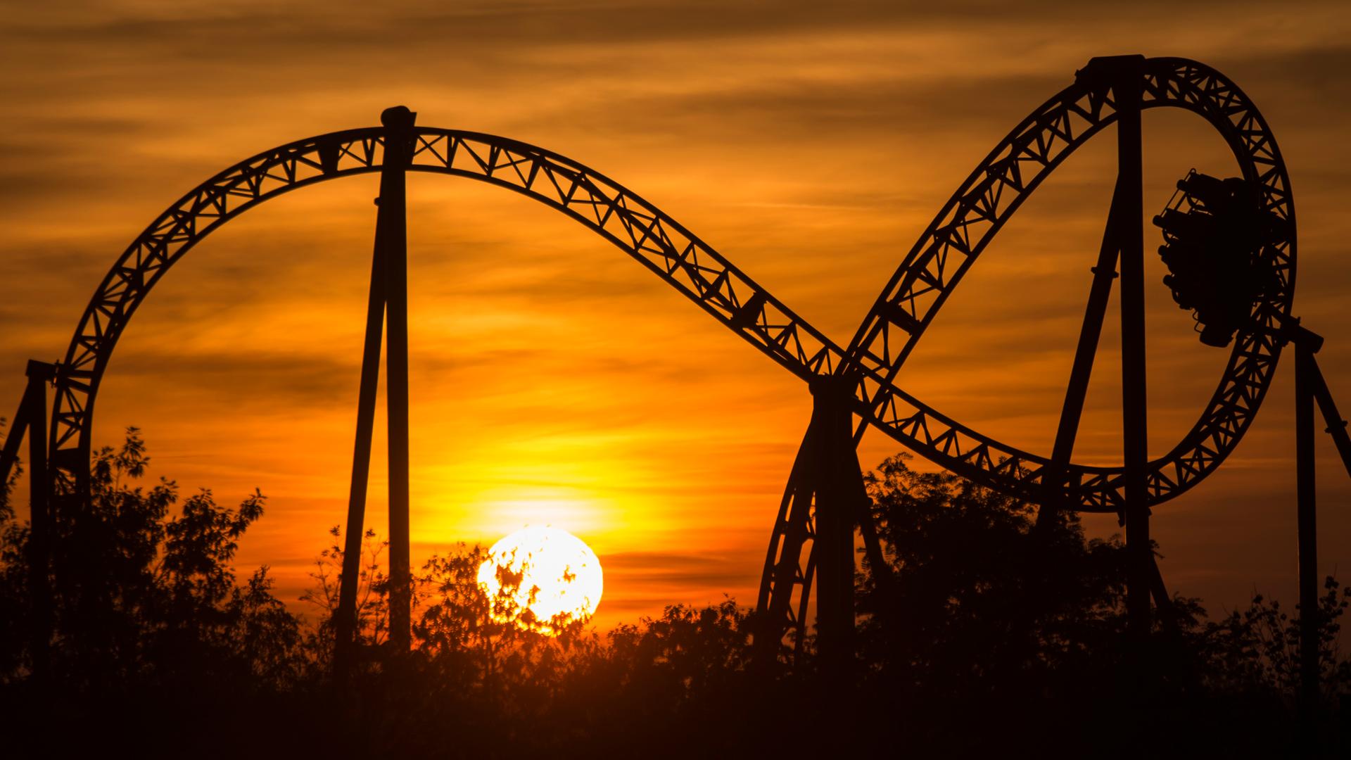 Gegen den goldenen Abendhimmel hebt sich eine Achterbahn mit einer großen Schlaufe ab, in der gerade ein Wagen unterwegs ist. Gegen den goldenen Abendhimmel hebt sich eine Achterbahn mit einer großen Schlaufe ab, in der gerade ein Wagen unterwegs ist.