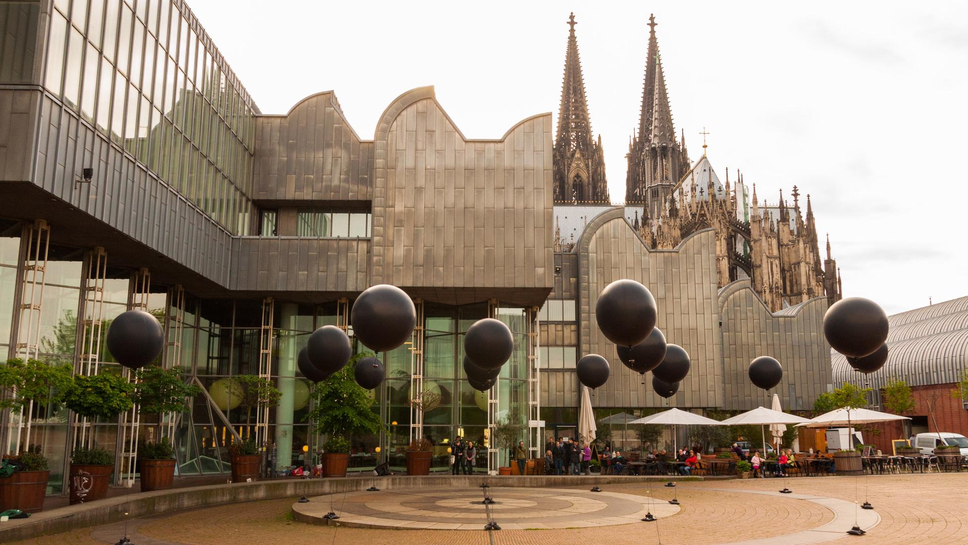 Vor der Kulisse von Philharmonie und Kölner Dom schweben sechzehn schwarze Ballons in der Luft, die eine musikalische Installation darstellen. Vor der Kulisse von Philharmonie und Kölner Dom schweben sechzehn schwarze Ballons in der Luft, die eine musikalische Installation darstellen.