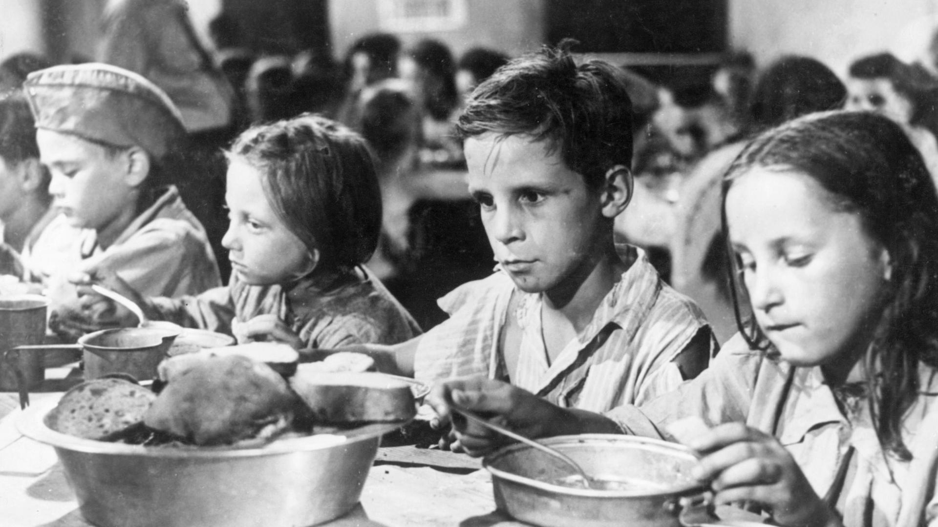 Mehrere Kinder essen auf dem Schwarz-Weiß-Bild an einem Tisch aus Blechtellern. Sie wirken ausgemergelt, die Augen eines Jungen sind leer. Mehrere Kinder essen auf dem Schwarz-Weiß-Bild an einem Tisch aus Blechtellern. Sie wirken ausgemergelt, die Augen eines Jungen sind leer.