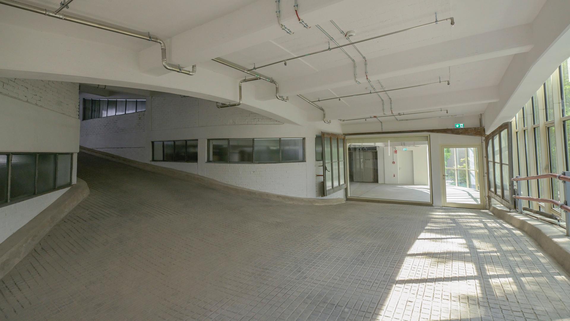 Das Bild zeigt eine Ebene eines ehemaligen Parkhauses, der Kant-Garagen in Berlin. Die Auffahrt ist zu erkennen, rechts ist eine Glasfront zu sehen, durch die das Laubwerk von Bäumen draußen zu erkennen ist. Der Raum ist geweißt und lichtdurchflutet. Das Bild zeigt eine Ebene eines ehemaligen Parkhauses, der Kant-Garagen in Berlin. Die Auffahrt ist zu erkennen, rechts ist eine Glasfront zu sehen, durch die das Laubwerk von Bäumen draußen zu erkennen ist. Der Raum ist geweißt und lichtdurchflutet.