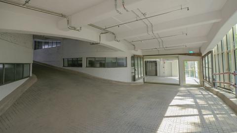 Das Bild zeigt eine Ebene eines ehemaligen Parkhauses, der Kant-Garagen in Berlin. Die Auffahrt ist zu erkennen, rechts ist eine Glasfront zu sehen, durch die das Laubwerk von Bäumen draußen zu erkennen ist. Der Raum ist geweißt und lichtdurchflutet.