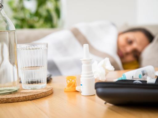 Eine Frau liegt krank auf einem Sofa (gestellte Szene), Medikamente liegen auf dem Tisch vor ihr