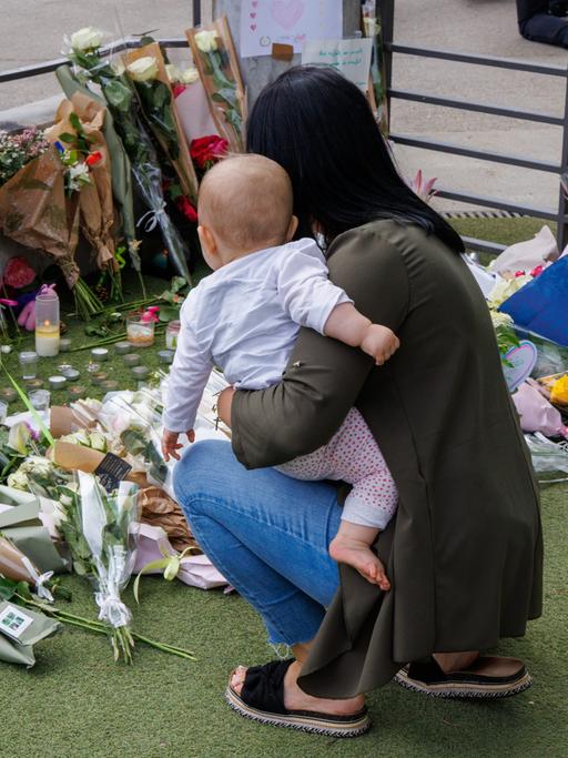 Eine Frau ist mit einem Kleinkind auf dem Arm in die Hocke gegangen und gedenkt der Opfer des Anschlags in der französischen Stadt Annecy. Auf dem BOden liegen Blumen, Stofftiere und ein LUftballon in Herzform. 