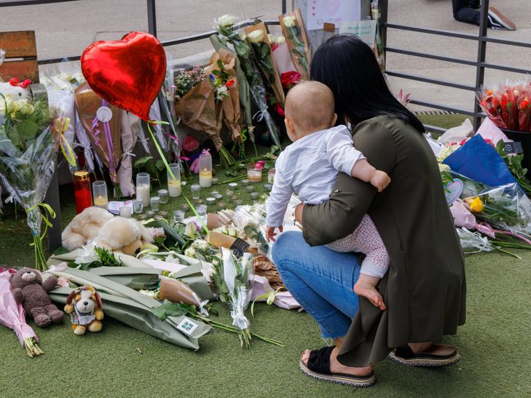 Eine Frau ist mit einem Kleinkind auf dem Arm in die Hocke gegangen und gedenkt der Opfer des Anschlags in der französischen Stadt Annecy. Auf dem BOden liegen Blumen, Stofftiere und ein LUftballon in Herzform. 