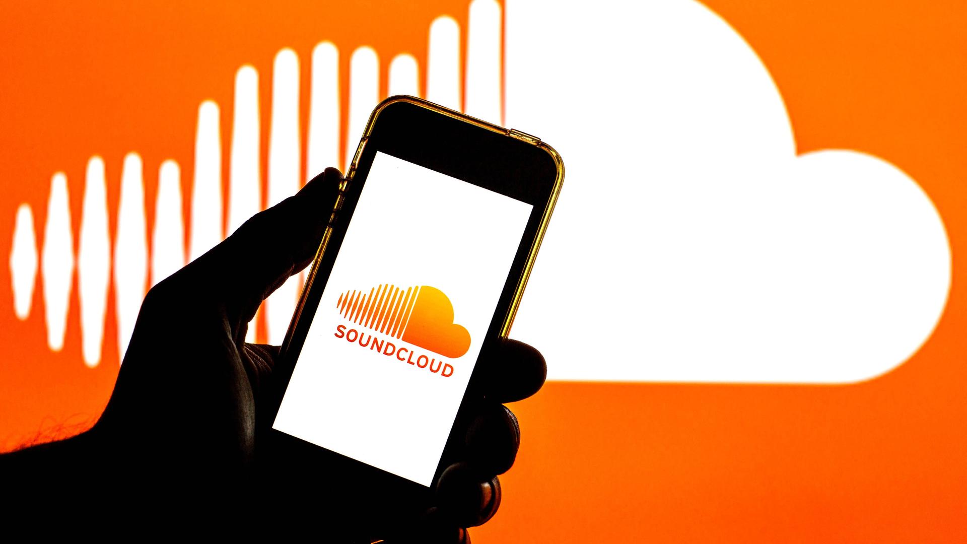 In der Illustration ist eine Hand zu sehen, die ein Smartphone hält. Auf dem Bildschirm ist das Soundcloud-Logo, auch im Hintergrund ist das Logo, eine weiße Wolke auf orangem Grund, wobei die Wolke links gestrichelt ist. In der Illustration ist eine Hand zu sehen, die ein Smartphone hält. Auf dem Bildschirm ist das Soundcloud-Logo, auch im Hintergrund ist das Logo, eine weiße Wolke auf orangem Grund, wobei die Wolke links gestrichelt ist.