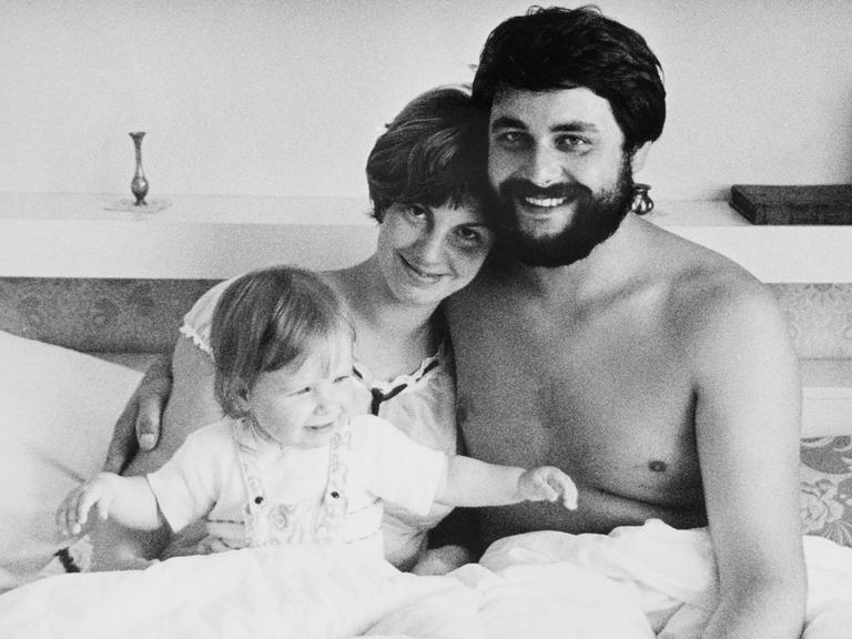 Schwarzweiss Aufnahme von einem jungen Paar mit Baby im Bett, die Eltern lächeln glücklich in die Kamera. DDR, Leipzig 1978.